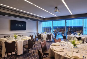 A conference room with multiple tables set for dining, a large screen displaying the hotel name, and a view of the cityscape through large windows. - Loews Chicago Hotel (Chicago)