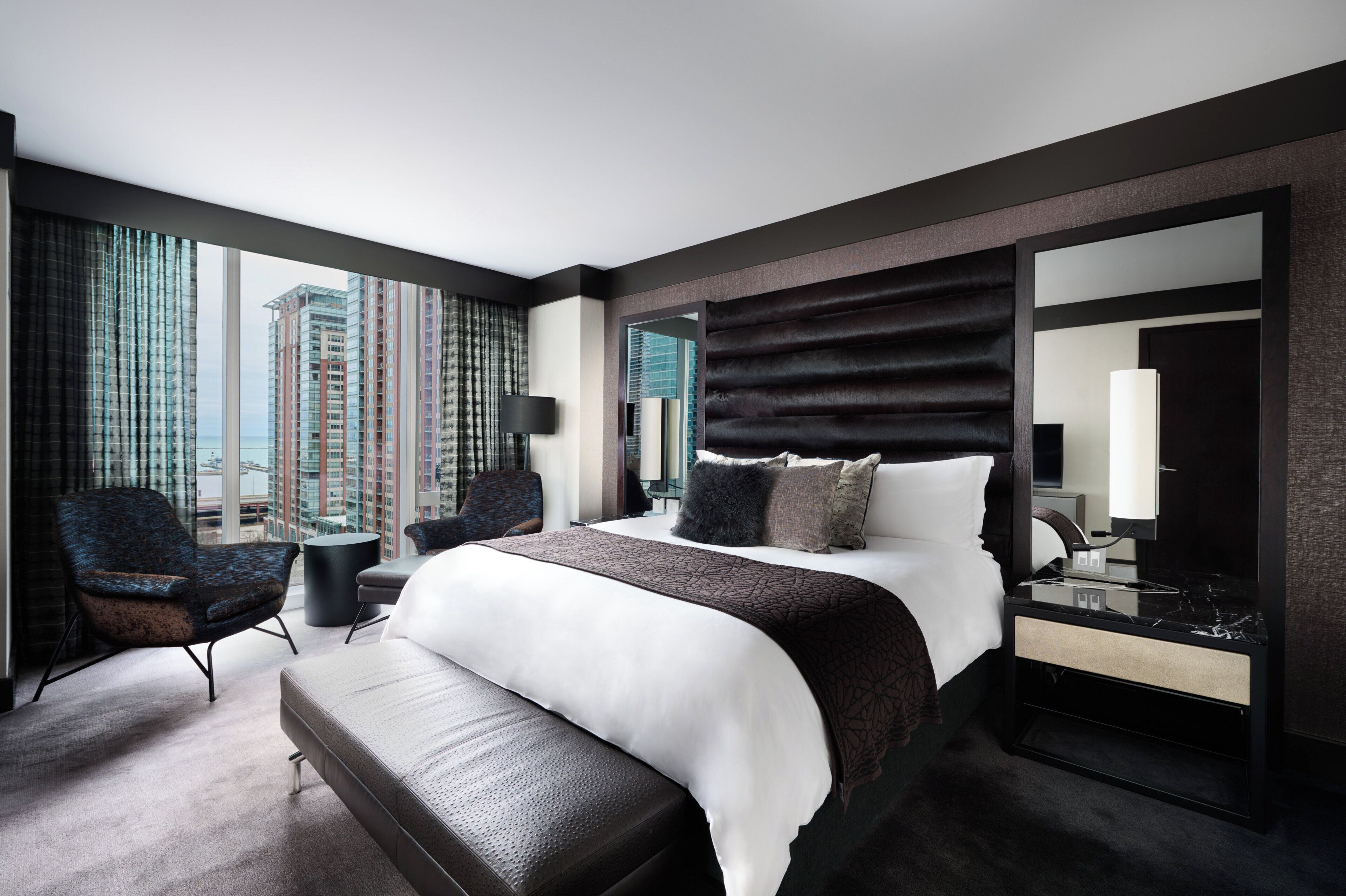 A modern hotel room with a large bed, a sitting area with a chair and a small table, a large window with a view of tall buildings, and a large mirror.