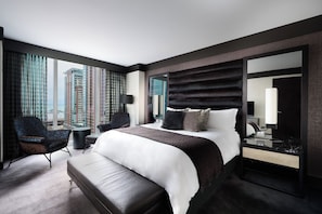 A modern hotel room with a large bed, a sitting area with a chair and a small table, a large window with a view of tall buildings, and a large mirror.