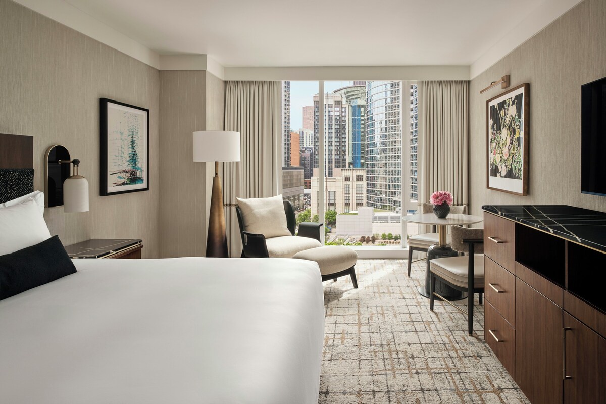 A hotel room with a bed, a desk, a chair, a lamp, and a view of the cityscape.