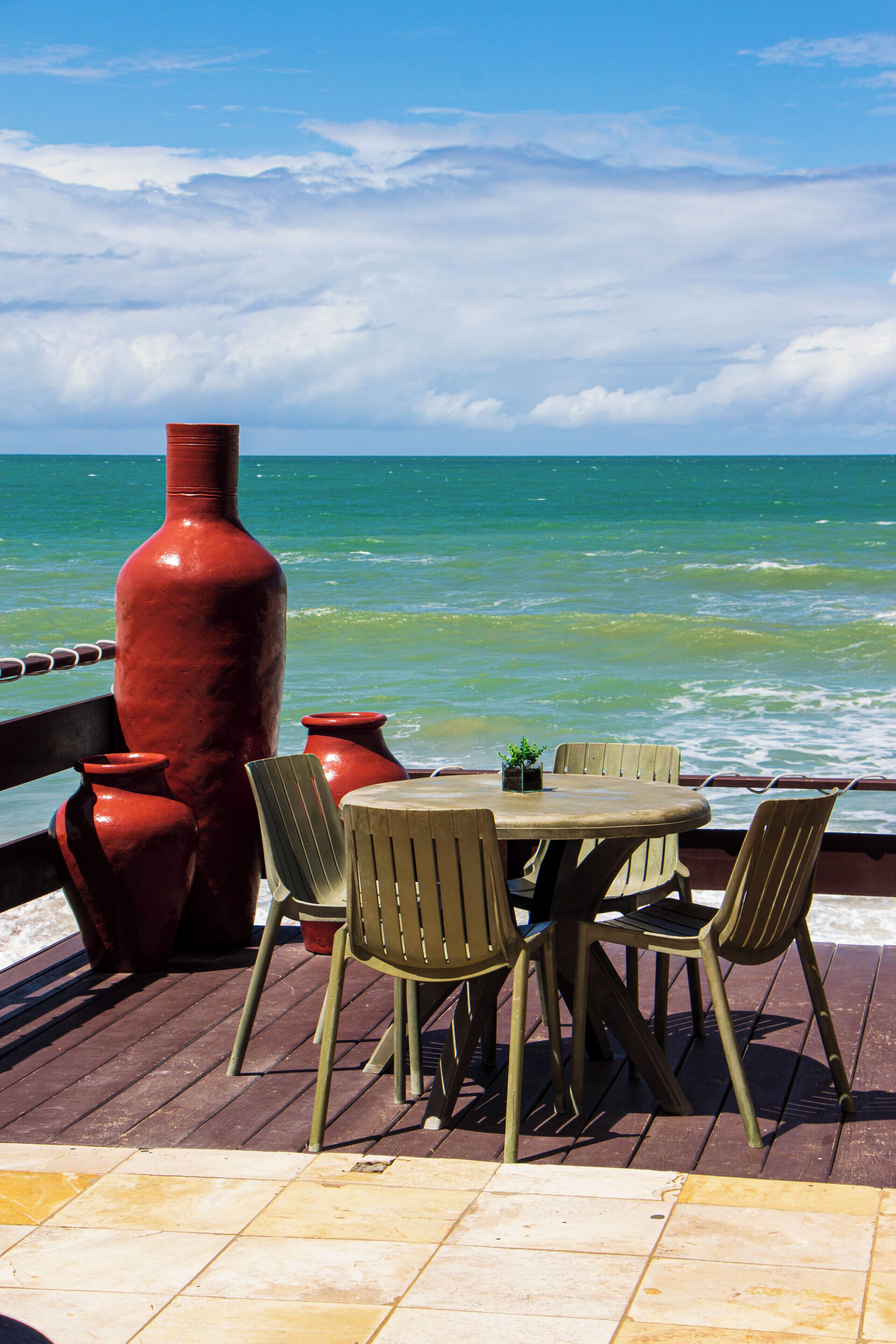 A wooden deck with a round table and chairs, large red vases, and a view of the ocean.
