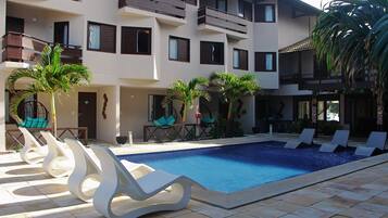 Una piscina con sillas de descanso, palmeras y un edificio de varios pisos con balcones.