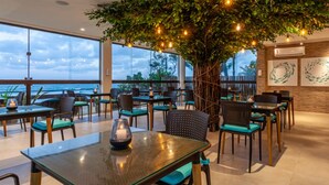 Un espace de restauration intérieur avec des tables et des chaises, un grand arbre décoré de guirlandes lumineuses et une vue sur l’océan grâce à de grandes fenêtres.
