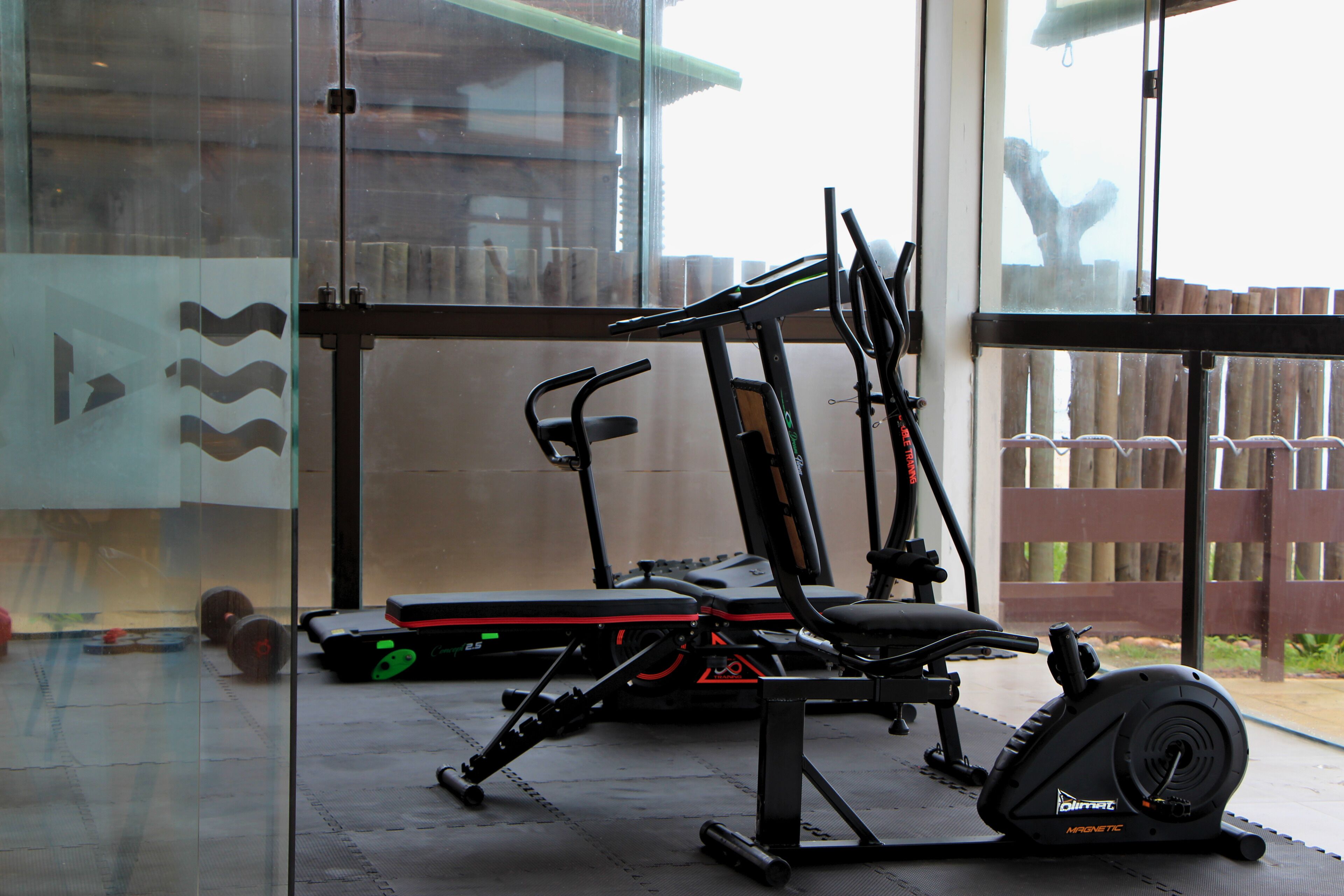 A gym with exercise equipment, a glass wall, and a wooden fence visible through the window.