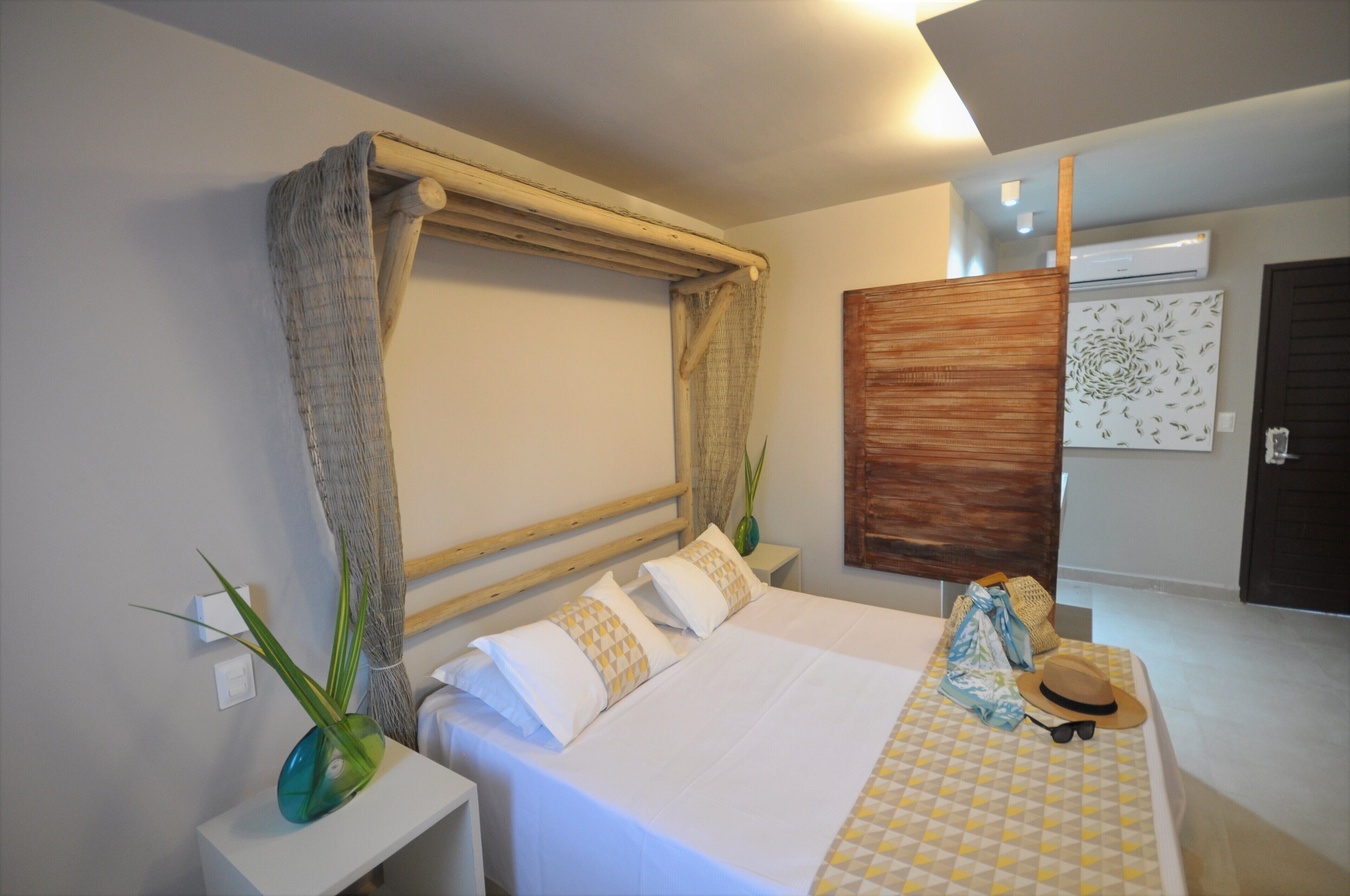 A hotel room with a bed, a nightstand, a vase with a plant, a hat, sunglasses, and a patterned tablecloth.