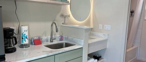 A modern kitchen with a built-in microwave, a coffee maker, and a sink with a mirror above it. There is a shelf with dishes and a towel rack.