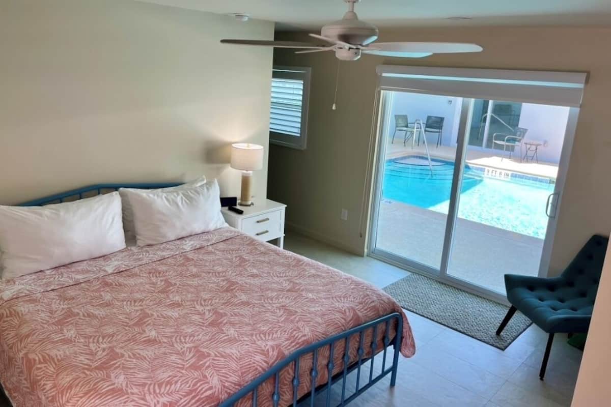 A bedroom with a bed, a ceiling fan, a nightstand, a lamp, and a view of a swimming pool through a sliding glass door.