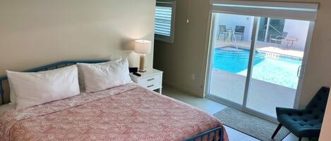 A bedroom with a bed, a ceiling fan, a nightstand, a lamp, and a view of a swimming pool through a sliding glass door.