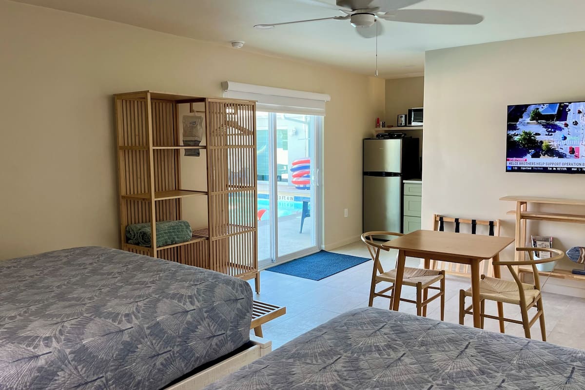 A room with two beds, a dining table with chairs, a ceiling fan, a TV, and a view of the outside.