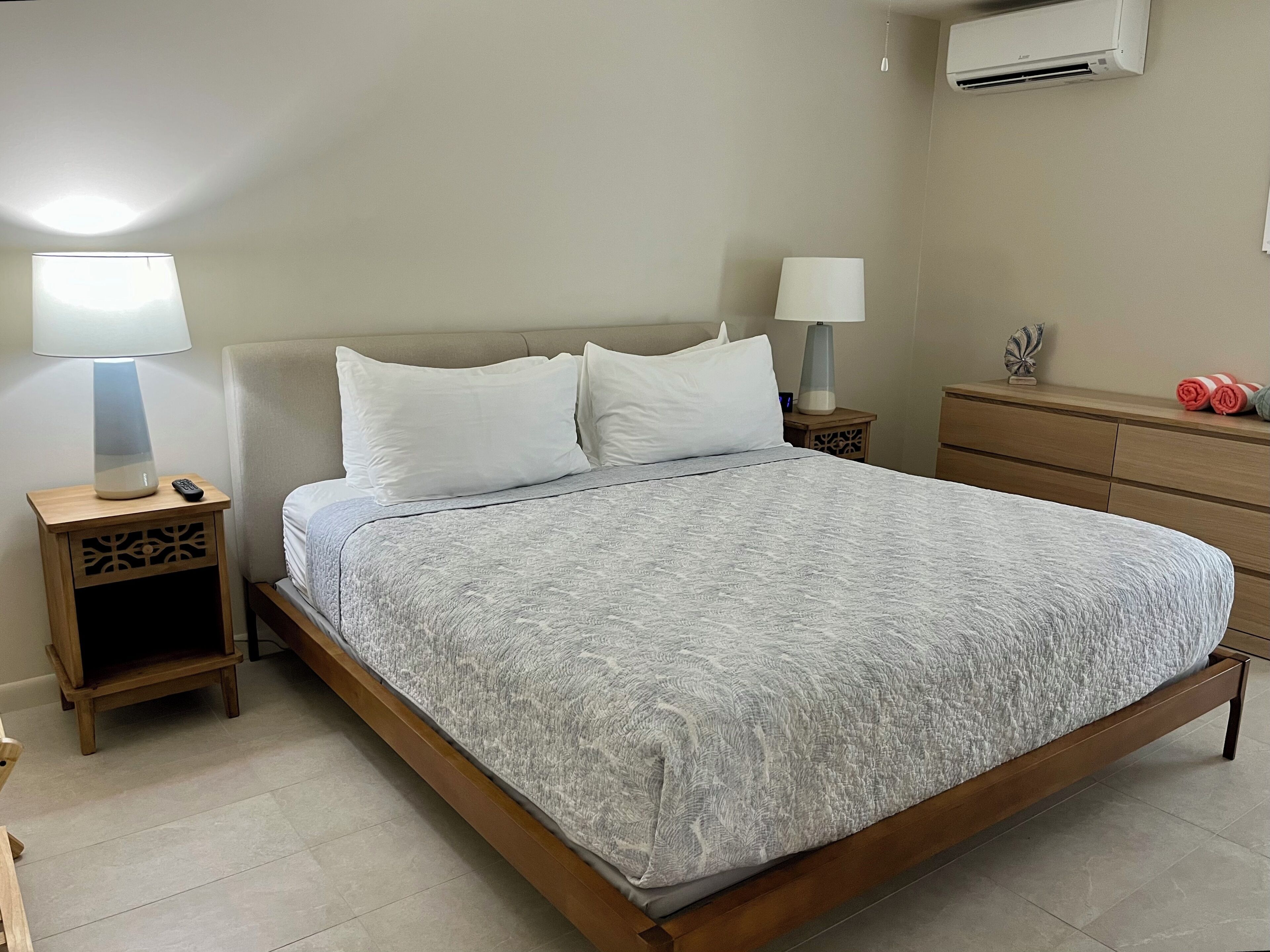 Een net opgemaakt bed met een gebloemd plaid, twee nachtkastjes met lampen, een houten dressoir en een airconditioner aan de muur.