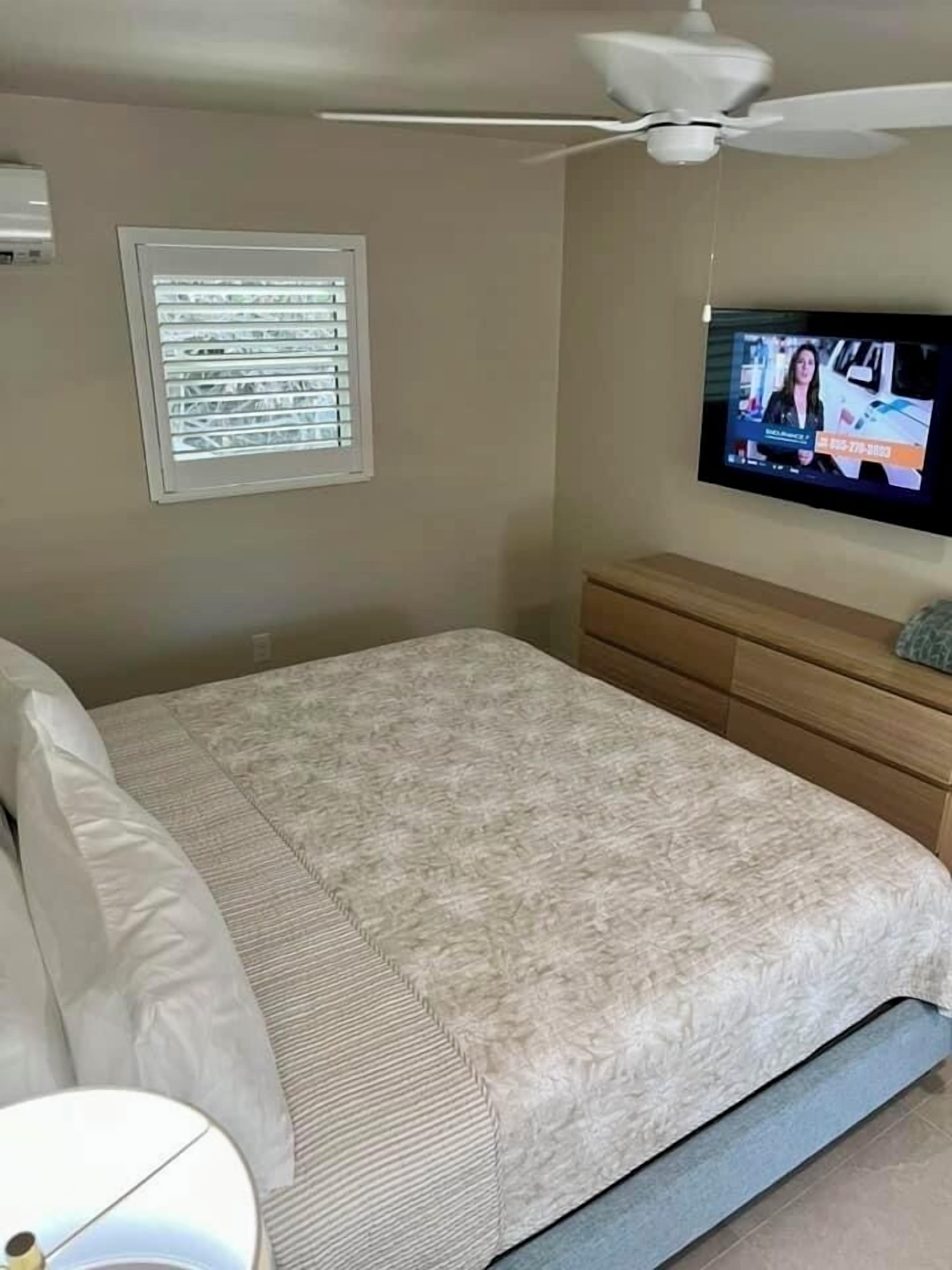 A bedroom with a bed, a ceiling fan, a window with blinds, a TV on the wall, and a wooden dresser.