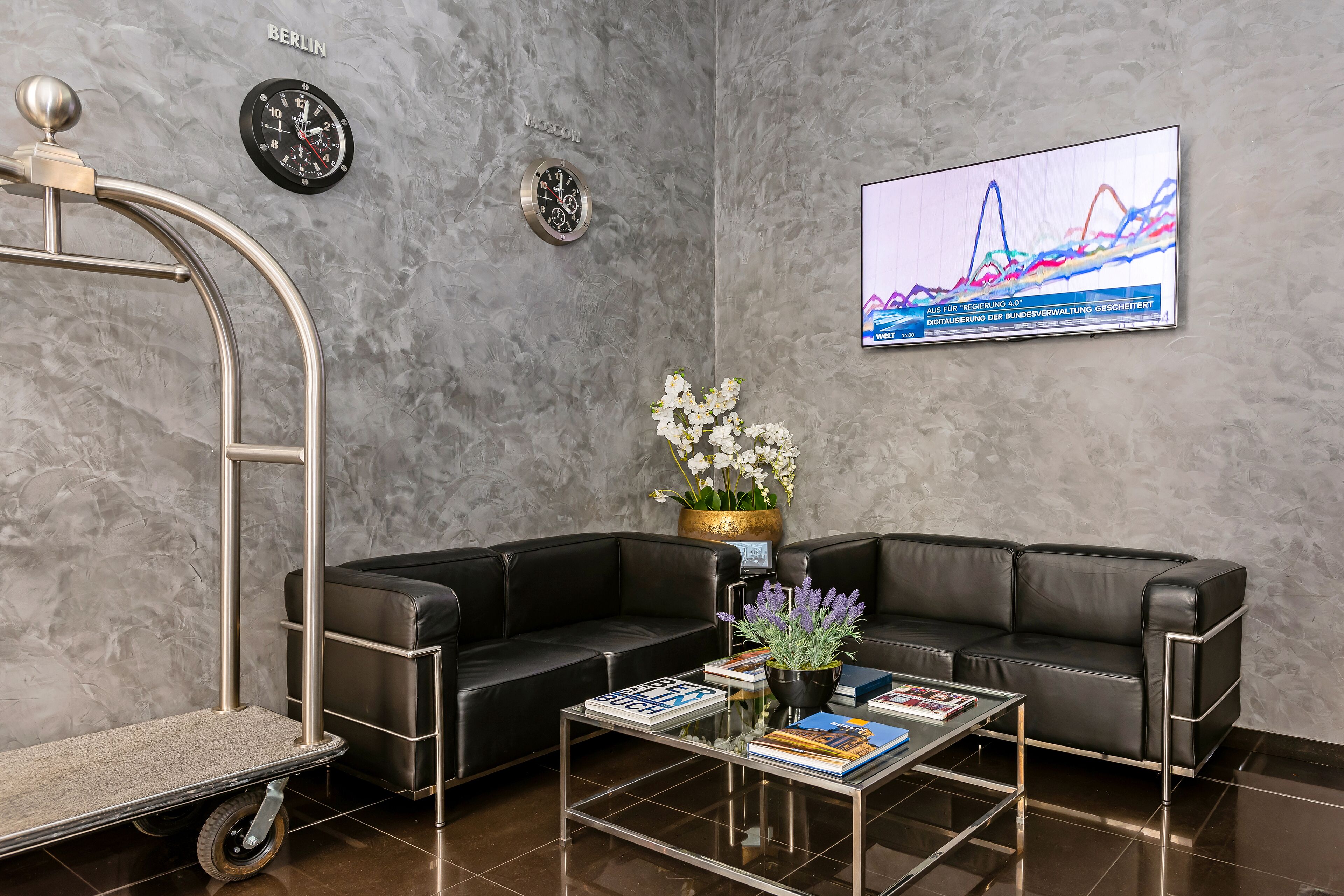 Un hall d’entrée moderne avec un canapé en cuir noir, une table basse en verre sur laquelle sont posés des livres, une horloge murale et un téléviseur affichant un graphique.