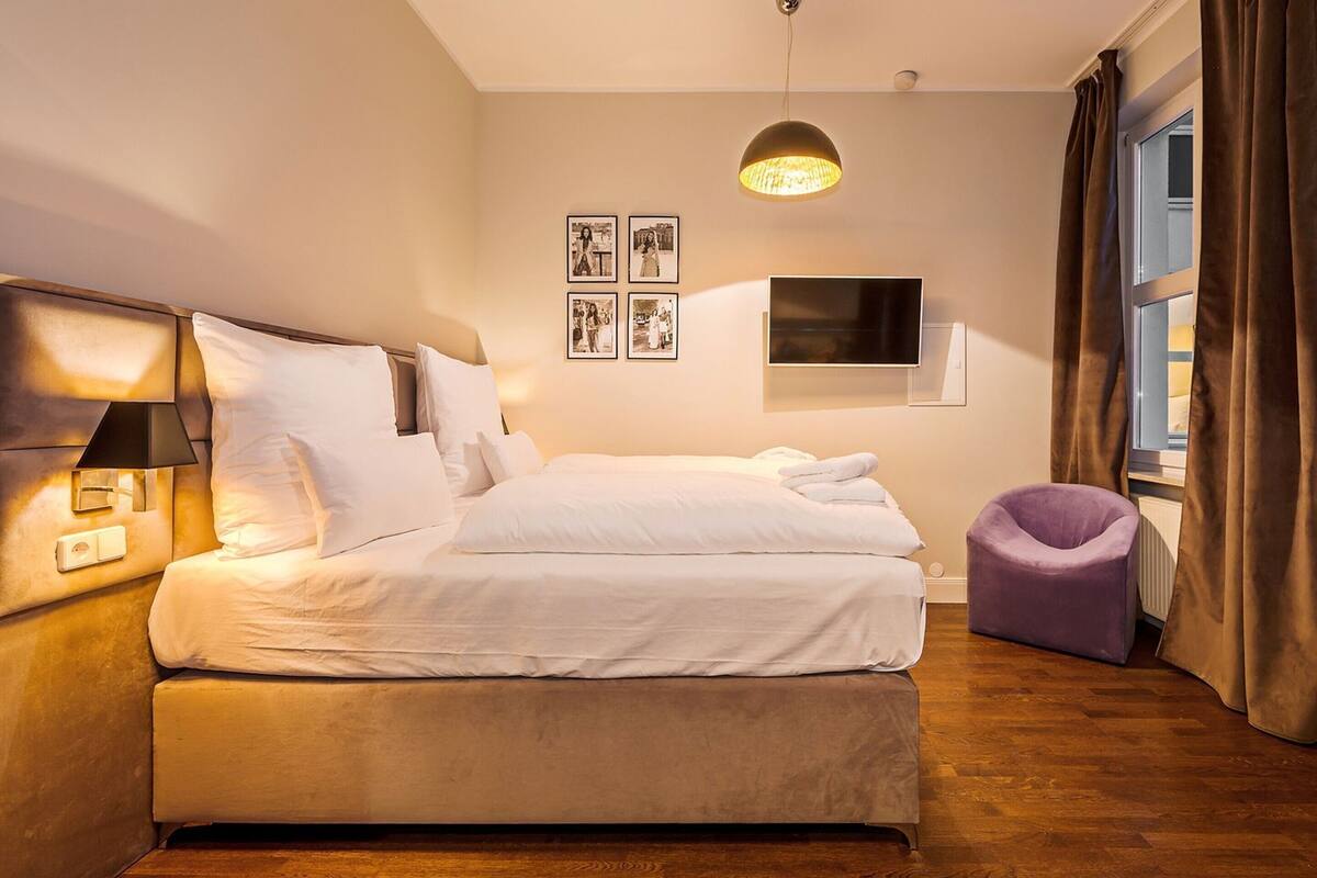 Habitación de hotel con cama, televisor, lámpara, silla y ventana con cortinas.