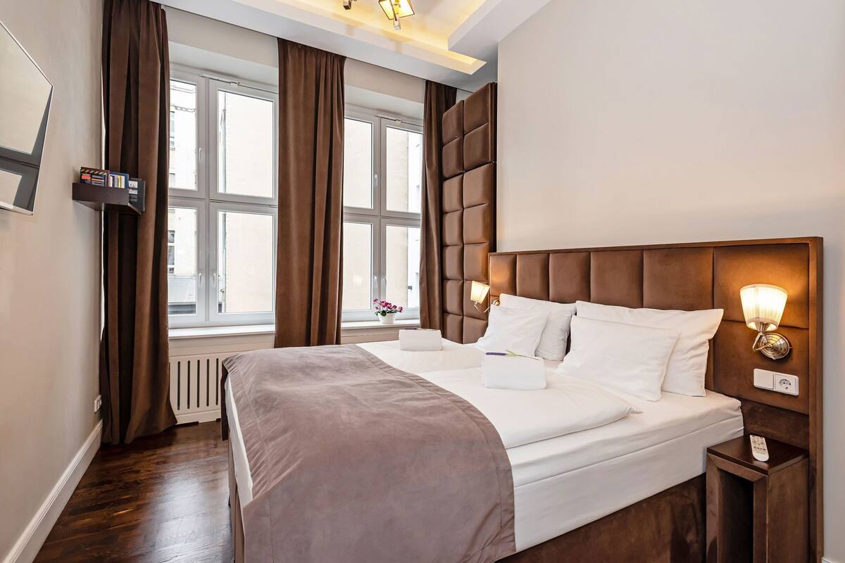Habitación de hotel con una cama grande, una mesita de noche con lámpara, una ventana con cortinas y un radiador.