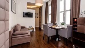 Ein modernes Wohnzimmer mit einer Couch, einem Esstisch mit Stühlen, einem an der Wand befestigten Fernseher und einem großen Fenster mit Vorhängen.