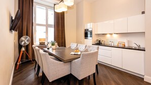 Eine moderne Küche mit weißen Schränken, ein Essbereich mit einem Holztisch und grauen Stühlen sowie ein Ventilator auf einem Stativ.