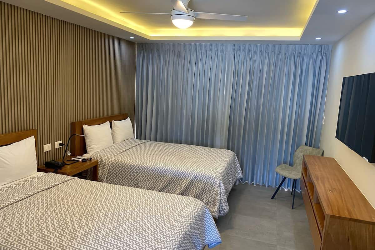A hotel room with two beds, a desk, a chair, a ceiling fan, and a large window with curtains.