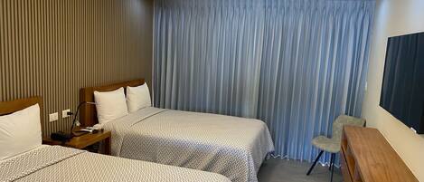 Habitación de hotel con dos camas, un escritorio, una silla, un ventilador de techo y un ventanal grande con cortinas.