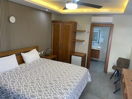 A modern hotel room with a large bed, wooden wardrobe, bedside table, and a small desk with a chair.