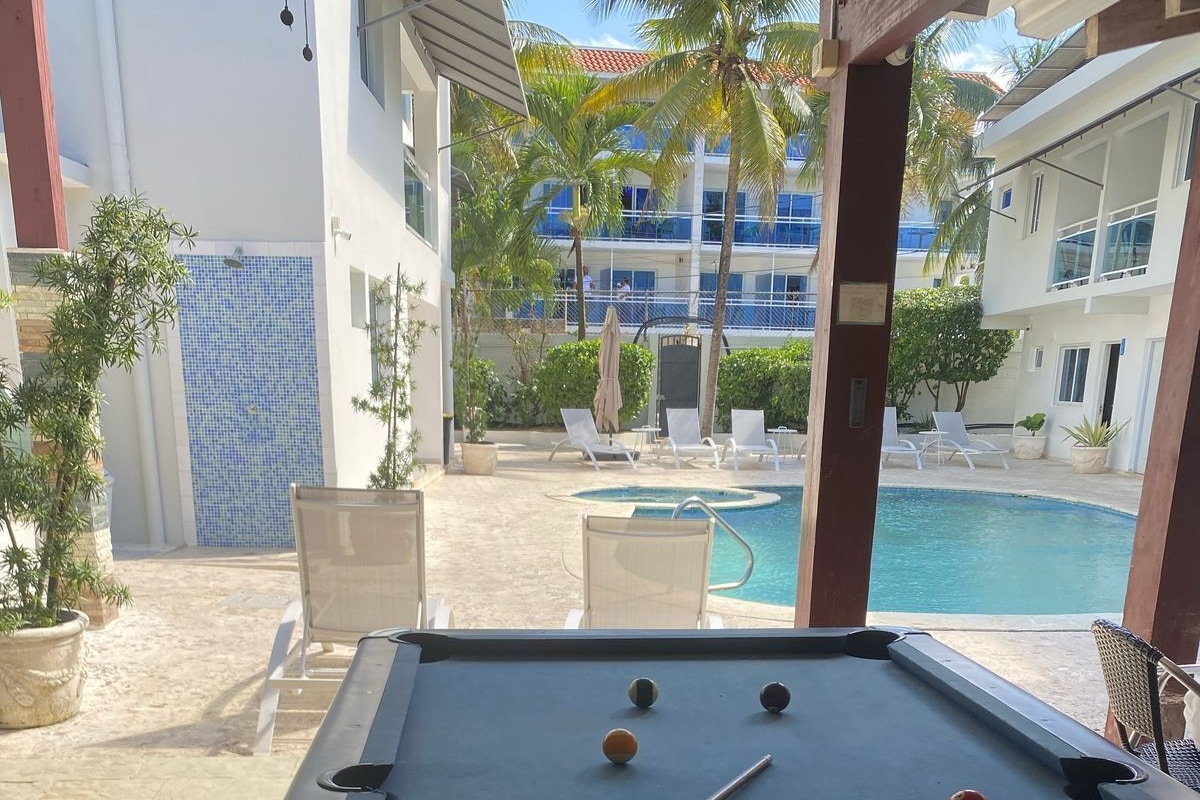 A pool table with balls and a pool cue in a covered outdoor area with a pool and lounge chairs.