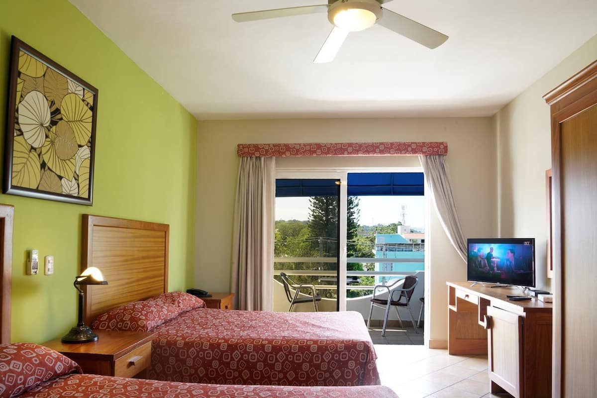 A hotel room with a bed, a desk with a television, a balcony with a view, and a ceiling fan.