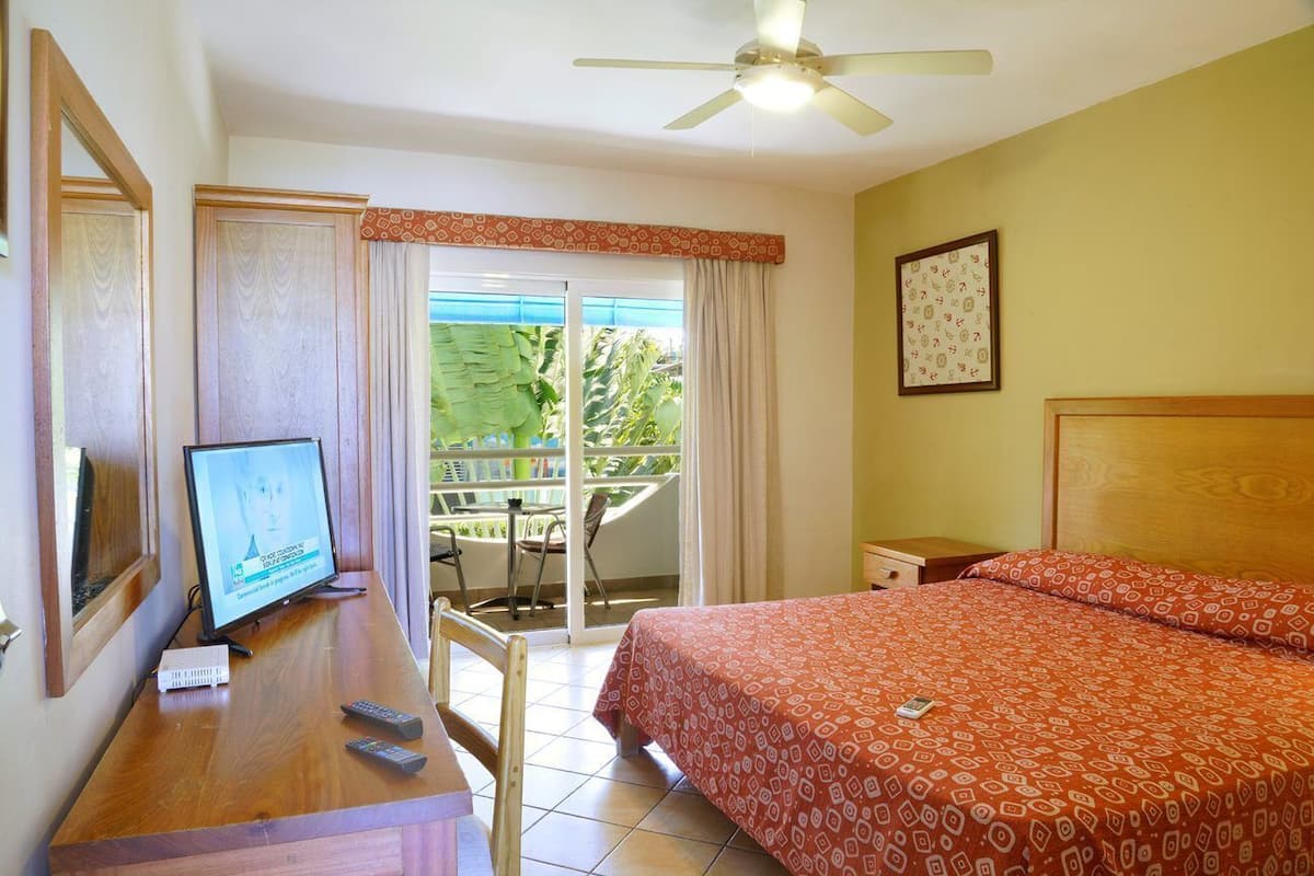 A hotel room with a bed, a desk with a computer, a ceiling fan, and a window with a view of greenery.