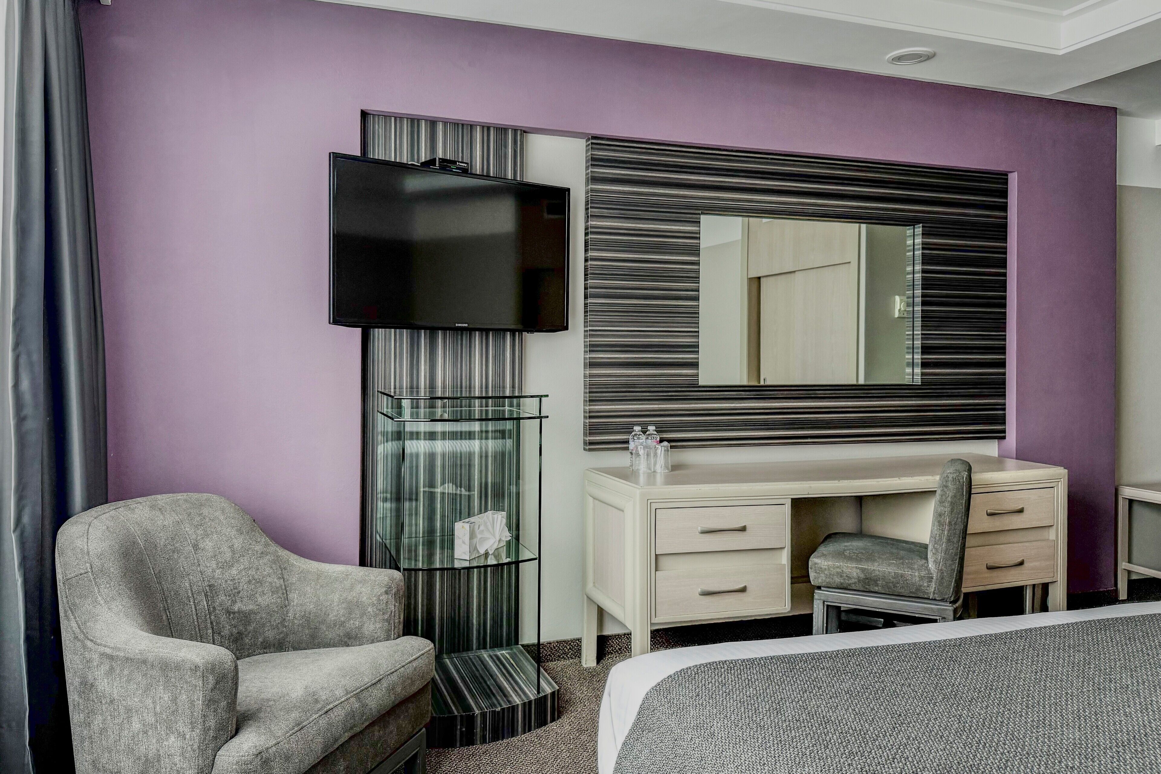 A hotel room with a bed, a TV, a desk with a chair, a glass shelf, and a mirror.