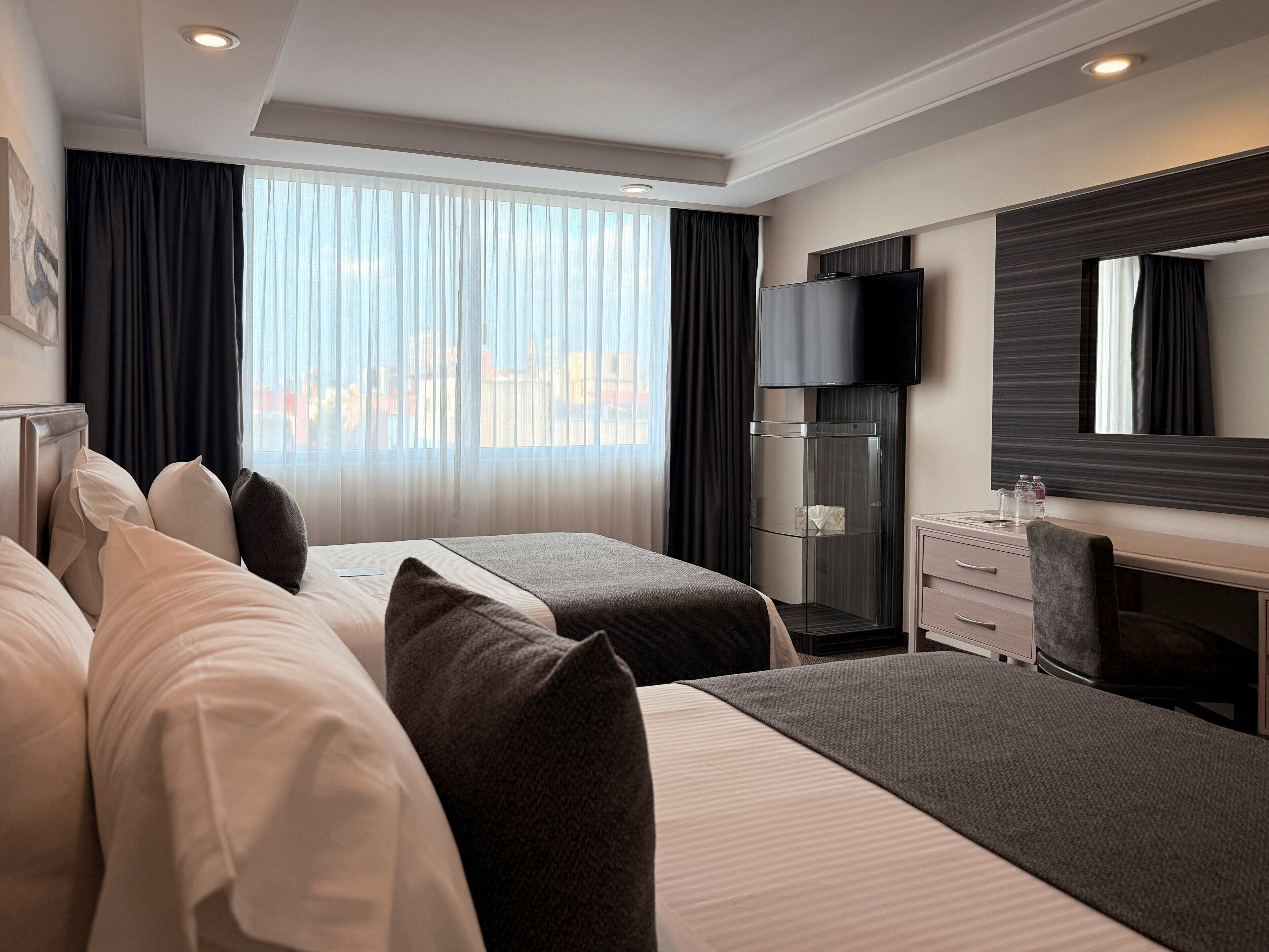 A hotel room with two beds, a desk, a TV, and a large window with curtains.
