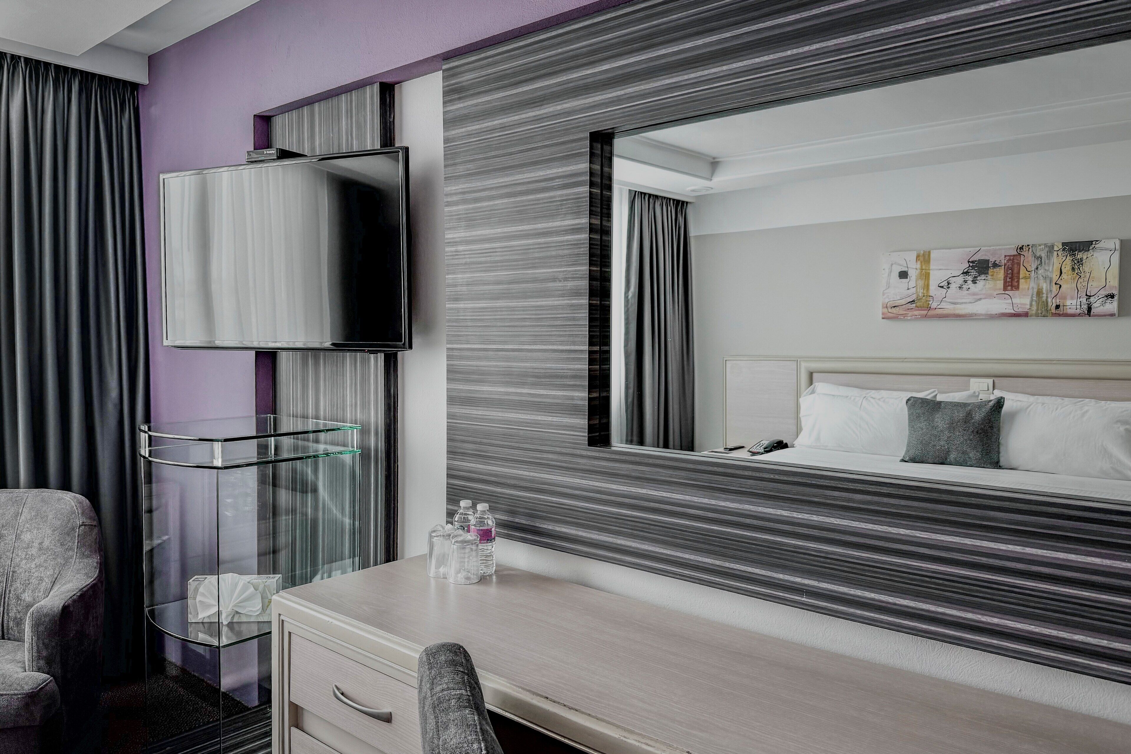 A hotel room with a bed, a TV, a glass side table, a chair, and a painting on the wall.