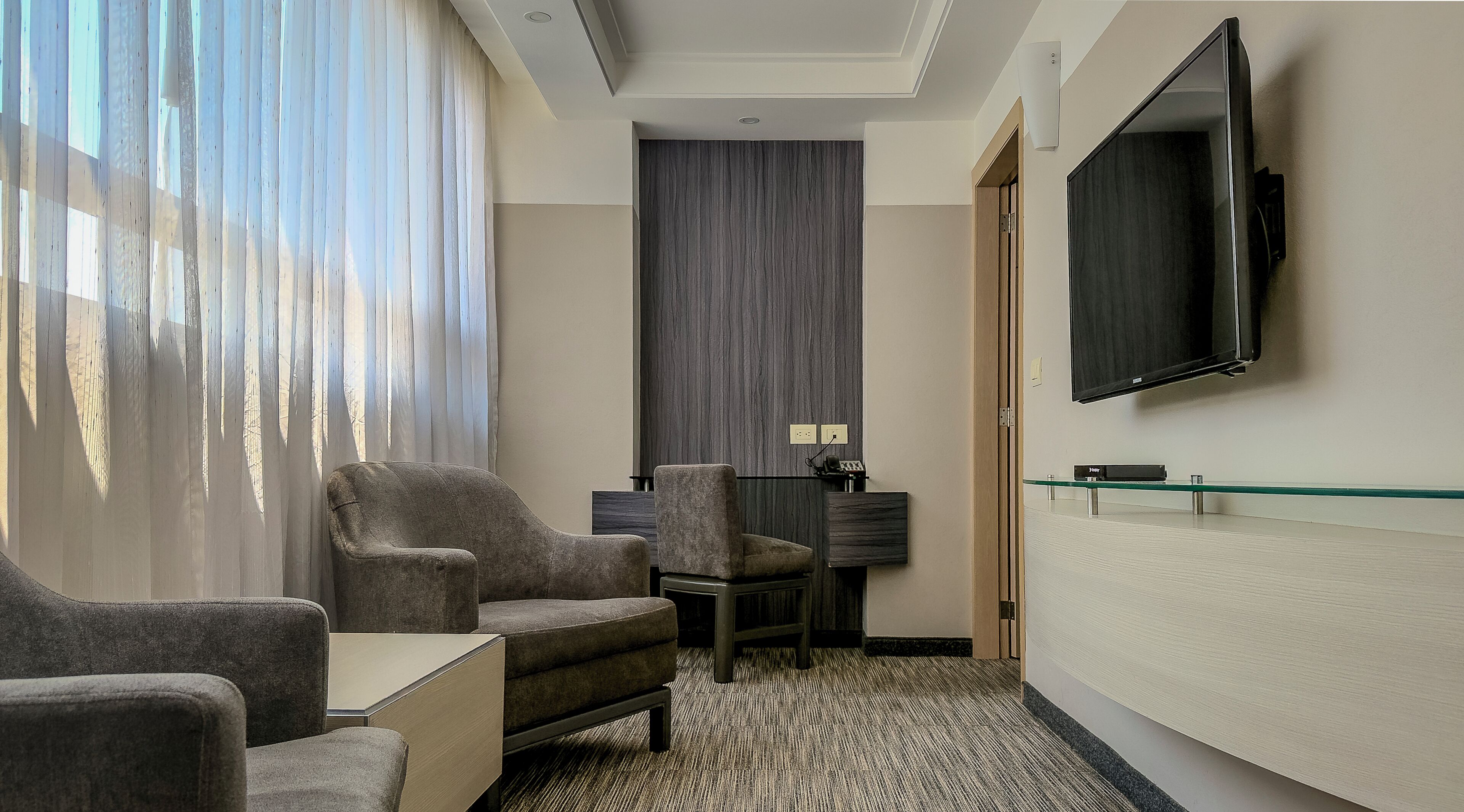 Hotelkamer met zithoek, bureau, flatscreen televisie en groot raam met gordijnen.