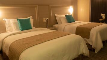 Habitación de hotel con dos camas, cada una con ropa blanca y almohadas color turquesa. Tiene un cabecero de madera, una lámpara de noche y un pequeño florero sobre la mesita de noche.