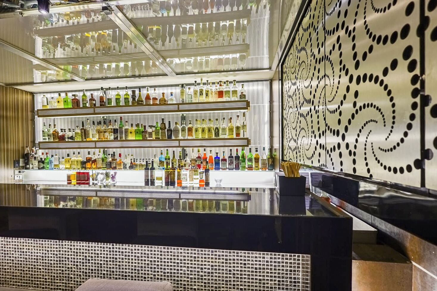 Zona de bar con estantes llenos de botellas de licores, un mostrador reflectante y un muro con diseño.