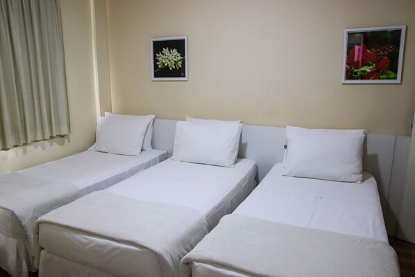 Habitación de hotel con tres camas individuales, cada una con ropa blanca y almohadas. Hay dos cuadros en la pared, uno con flores blancas y el otro mostrando una mariposa sobre flores rojas.