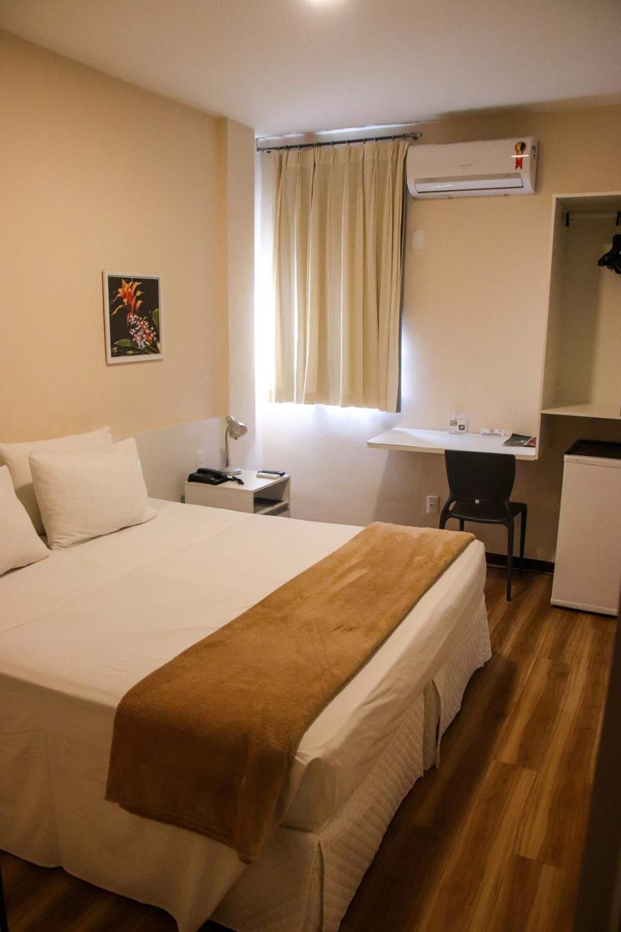 A hotel room with a large bed, a desk, a chair, an air conditioning unit, and a window with curtains.