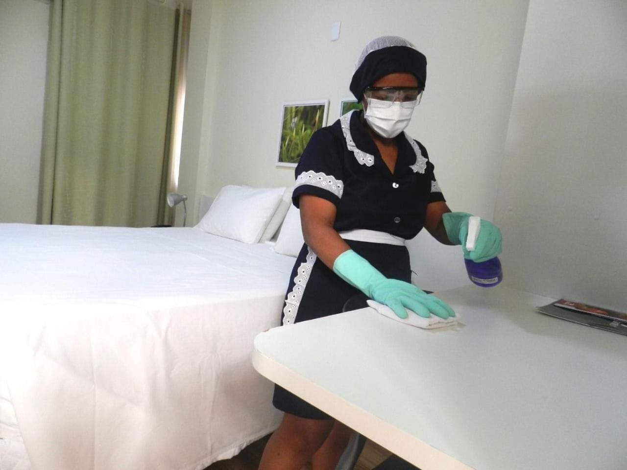 A hotel room with a bed, a bedside table, and a person cleaning the table.
