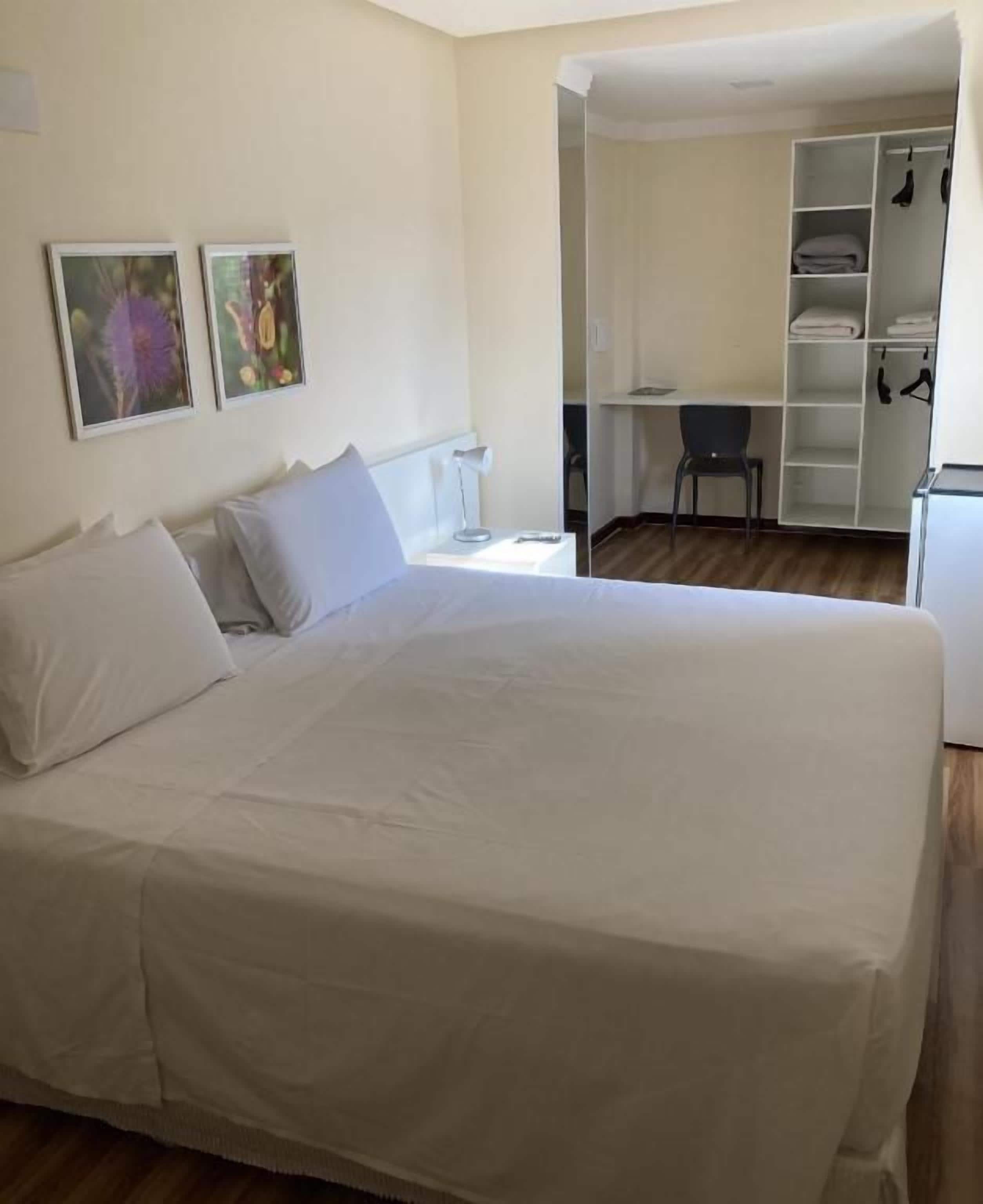 Ein Schlafzimmer mit einem Bett, einem Schreibtisch und einem Stuhl. Ein Kleiderschrank bietet Stauraum für Kleidung und es gibt ein Regal.