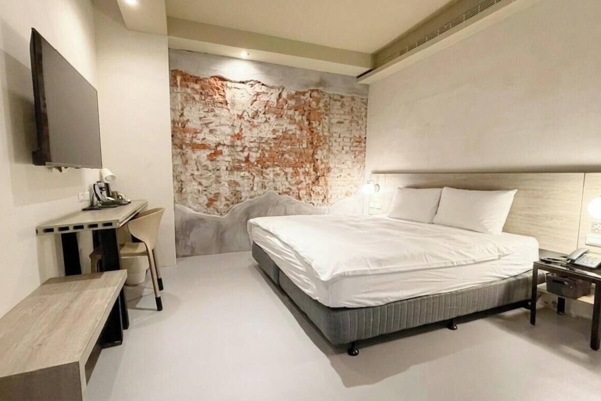 A modern hotel room with a bed, bedside table, chair, and a TV mounted on the wall.