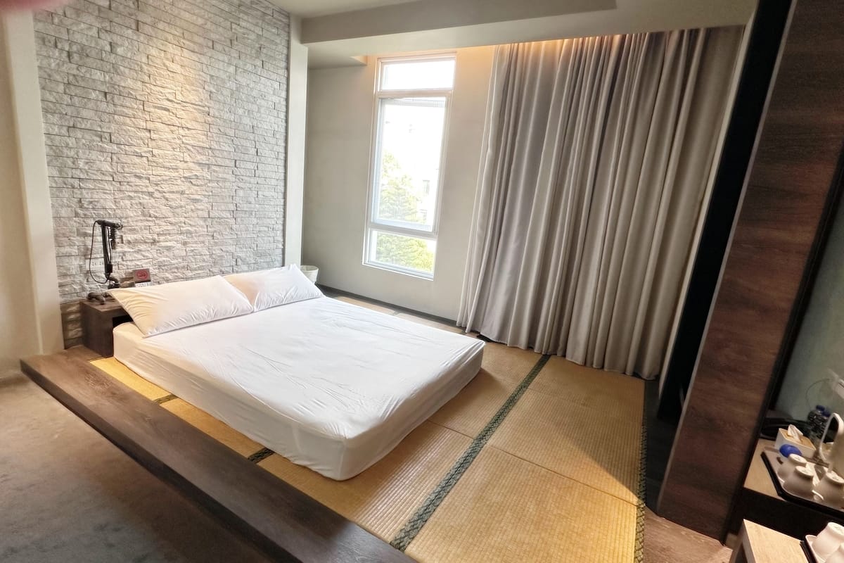 A hotel room with a large bed, a stone accent wall, a window with curtains, and a small bedside table with a lamp.