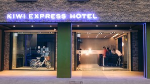 Exterior del hotel Kiwi Express con un letrero iluminado llamativo, una entrada de vidrio y una bicicleta estacionada afuera.