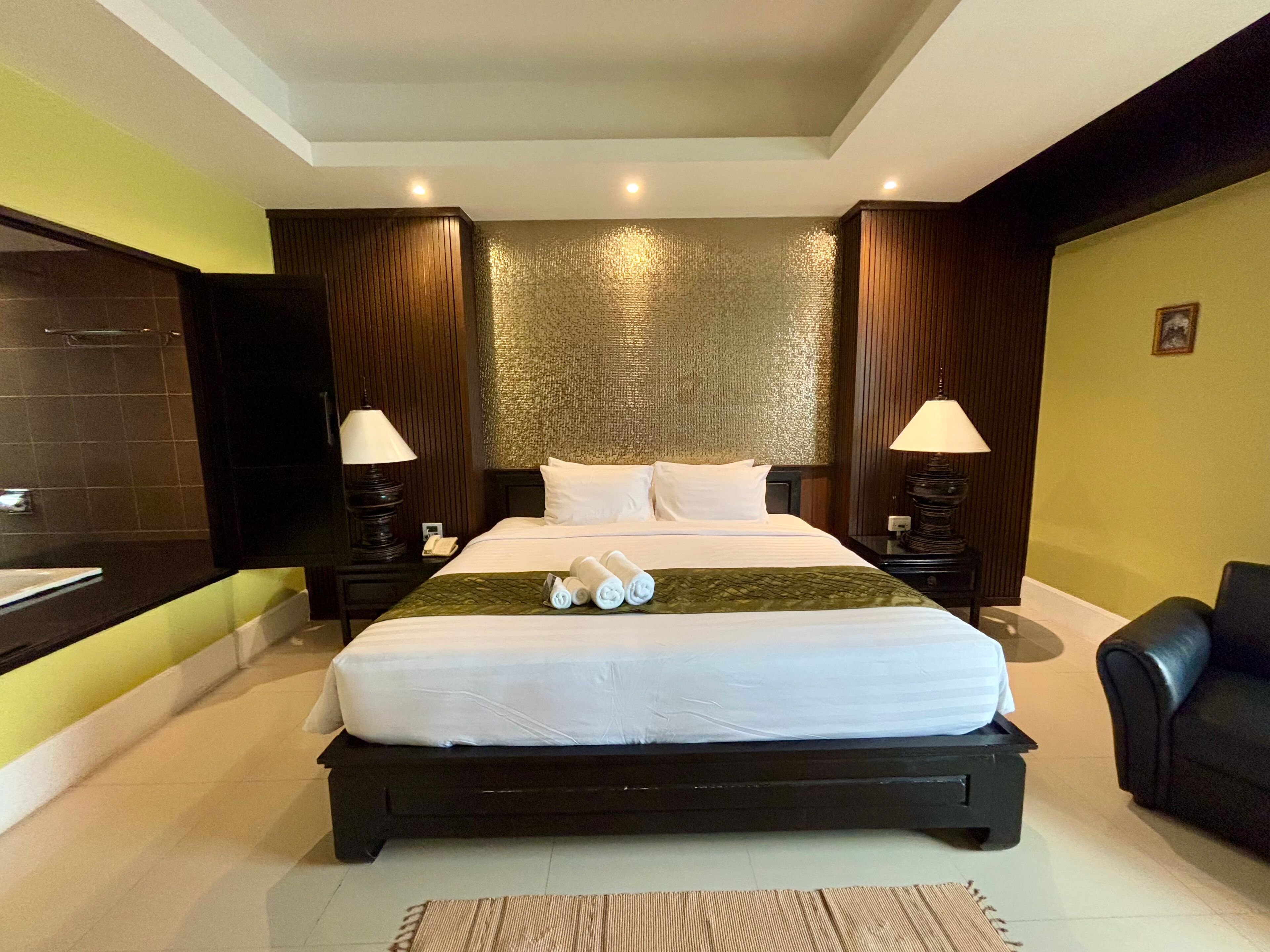 Habitación de hotel con una cama grande, dos mesitas de noche con lámparas, un cabecero de madera, un televisor montado en la pared y un baño con un espejo grande y un perchero para toallas.