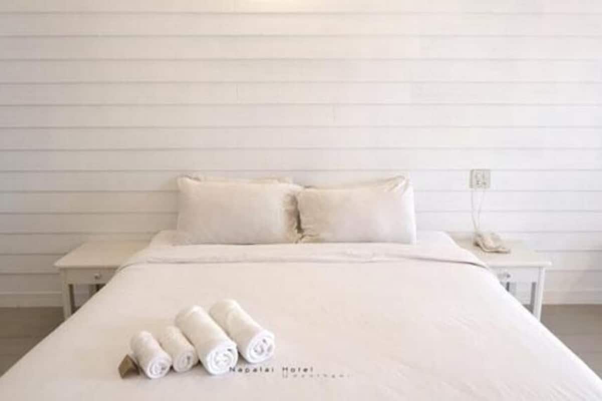 Una cama bien tendida con almohadas y toallas enrolladas, una mesita de noche con teléfono, y una pared con líneas horizontales.