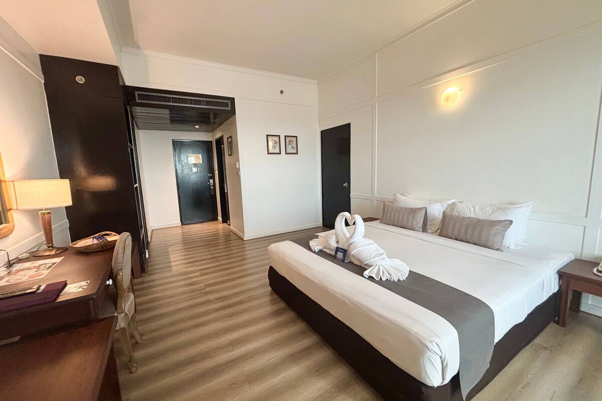 Habitación de hotel con una cama grande, un escritorio, una silla, una lámpara y una puerta que da a otra habitación.
