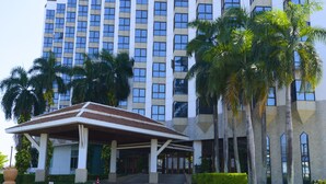 A modern hotel with a covered entrance, palm trees, and a large building in the background. - Napalai Hotel (Udon Thani)