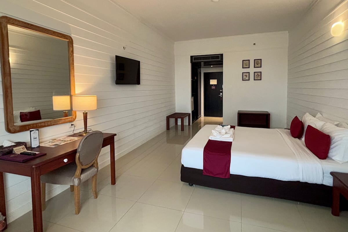 Habitación de hotel con cama, escritorio con silla, lámpara de escritorio, espejo grande, televisor y puerta que da a otra habitación.