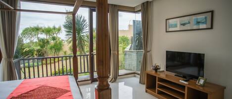 A bedroom with a large bed, a TV on a wooden stand, and a balcony with a view of greenery.