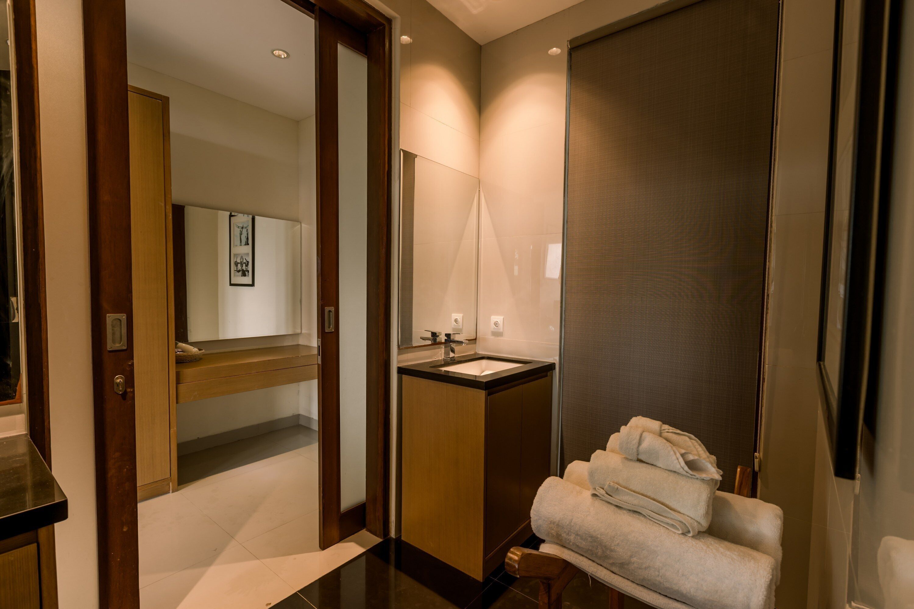 A modern bathroom with a wooden vanity, a sink, and a mirror. There is a towel rack with towels and a door leading to another room.