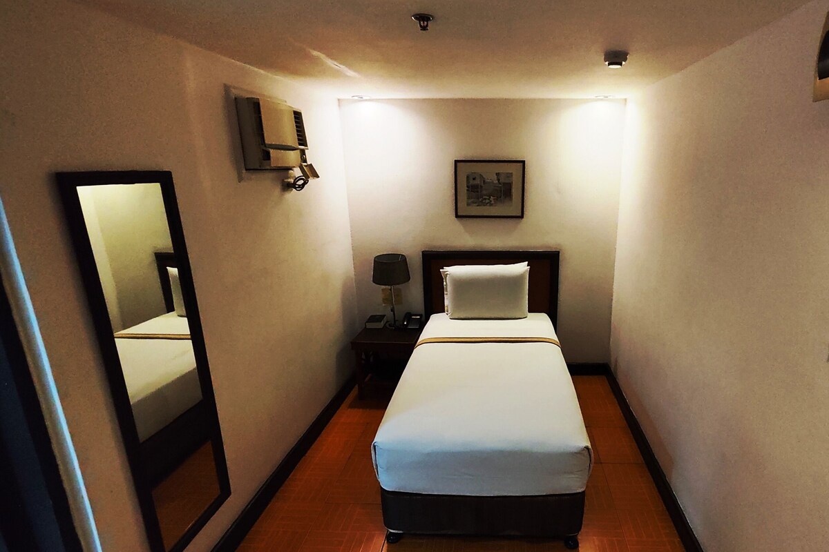Habitación de hotel estrecha con cama, mesita de noche, lámpara, espejo y lámpara de pared.