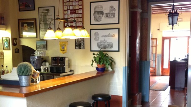 A restaurant interior with a bar area, stools, a table, and framed pictures on the walls. - Hotel Torino (Masnou)