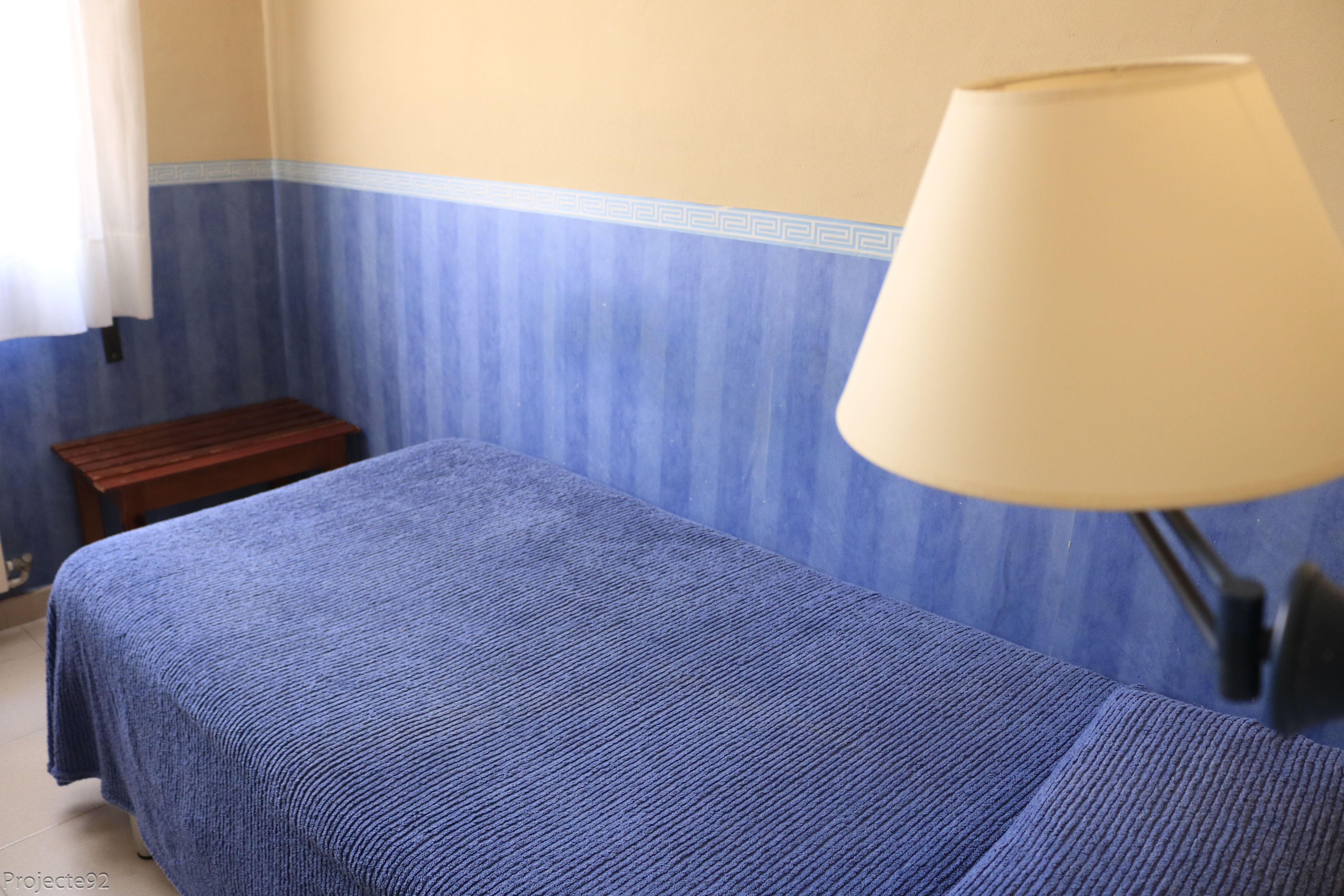 Ein Zimmer mit einem Bett, das mit einer blauen Decke bezogen ist, einem kleinen Holzschemel und einer Lampe mit einem beigen Lampenschirm.