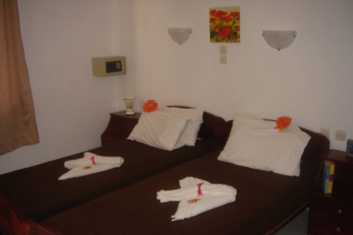Uma cama de casal com roupa de cama branca e flores laranjas, uma mesa de cabeceira com abajur e um aplique na parede.